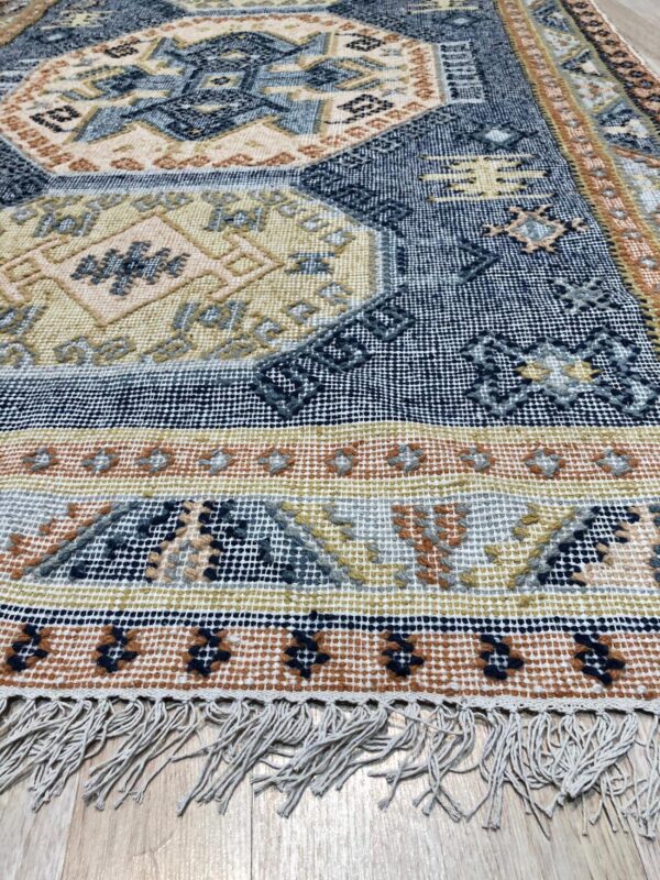 Geometric flatweave rug in blue, gold, and terracotta with white fringe on a wooden floor.