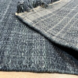 Textured blue-gray rug is folded on light wood flooring, revealing the woven backing and rolled fringe.