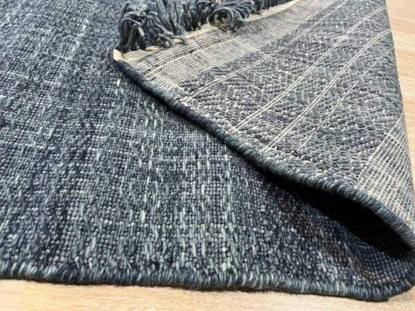 Textured blue-gray rug is folded on light wood flooring, revealing the woven backing and rolled fringe.