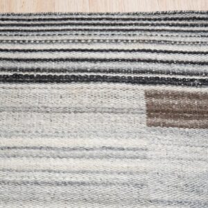 Flatweave rug with gray, black, and white stripes, featuring a brown square detail on light wood flooring.