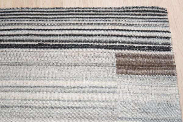 Flatweave rug with gray, black, and white stripes, featuring a brown square detail on light wood flooring.