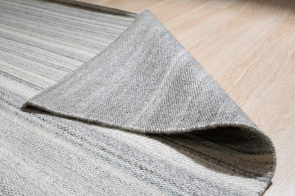 The edge of a gray and white striped flatweave rug is folded over a light wood floor.
