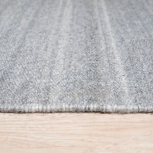 The bound edge of a textured, heathered light gray rug rests on a light blonde wood floor.