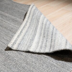 A monochromatic gray rug is flipped to show its striped backing on light wood plank flooring.