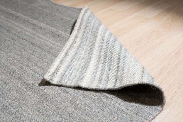 A monochromatic gray rug is flipped to show its striped backing on light wood plank flooring.