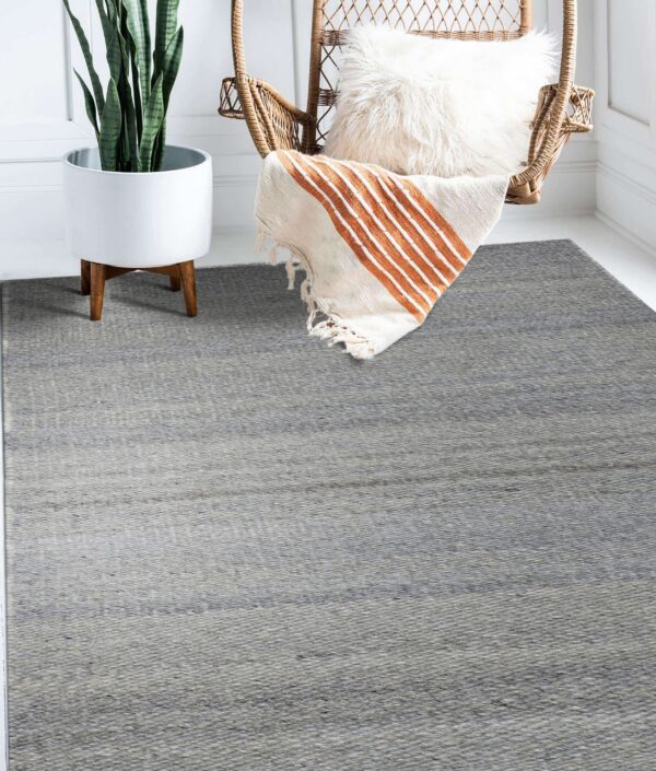 A flatwoven gray heathered rug is placed under a wicker chair and white plant stand on a white floor.