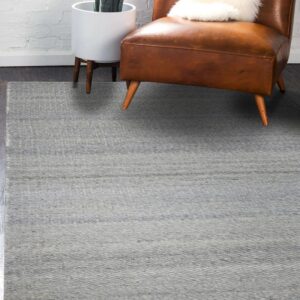 Light gray textured flatweave rug on dark wood flooring beside a brown leather chair and white brick wall.