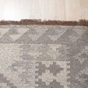 Beige and gray textured rug with a geometric pattern and dark brown fringed edge on light wood.