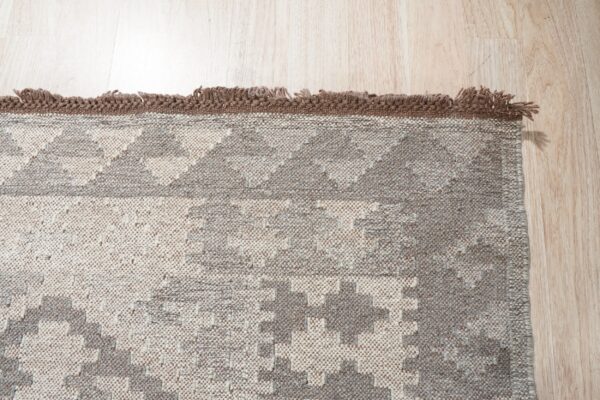 Beige and gray textured rug with a geometric pattern and dark brown fringed edge on light wood.
