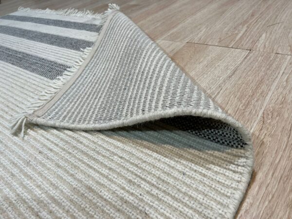Ribbed white and gray striped rug with fringe is pulled up on light wood flooring, showing the flat backing.