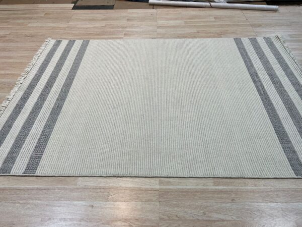 Ribbed cream rug with dark gray striped borders and visible fringe on light wood-look flooring.