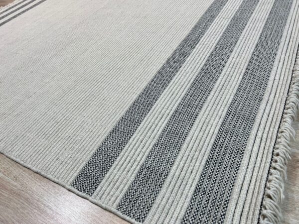Ribbed light-colored rug with thick black and white stripes and visible fringe on wood floor.