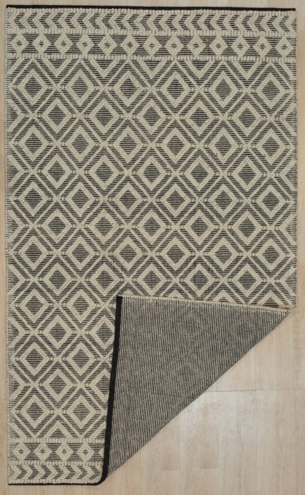 Geometric cream and dark gray rug featuring a diamond pattern, folded back to reveal the striped reverse side on wood flooring.