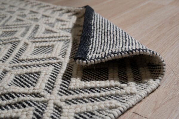 Cream and black textured diamond rug, partially rolled, rests on light beige wood flooring.