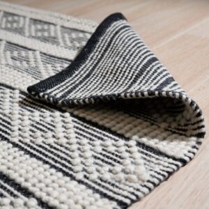 Black and off-white textured rug with geometric patterns, partially rolled on light wood floor.