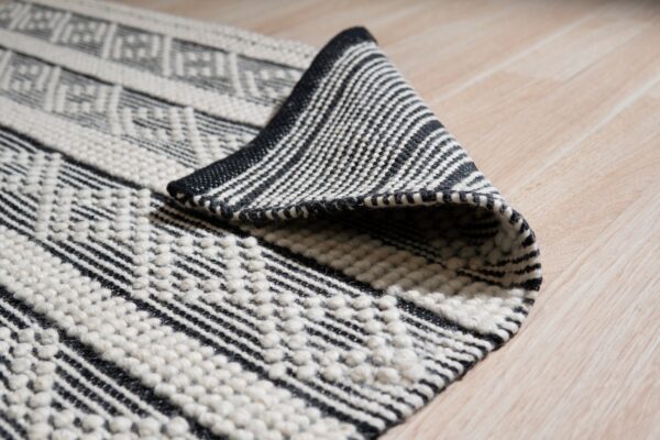 Black and off-white textured rug with geometric patterns, partially rolled on light wood floor.