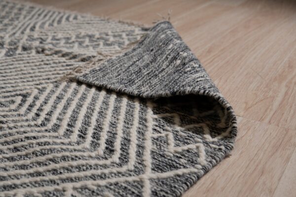 Close-up of a curled gray and white geometric textured rug on light wood flooring.
