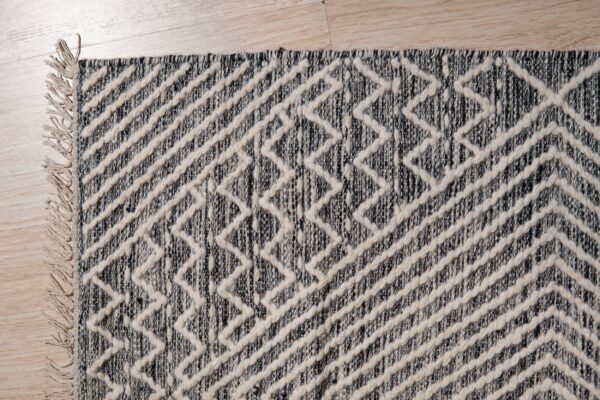 Raised cream geometric zigzags overlay a dark speckled rug with fringe on light wood flooring.