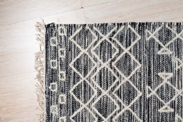 Black and off-white woven rug with raised diamond patterns and tassels resting on light wood planks.