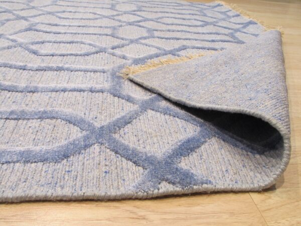 Light blue geometric rug with raised pile pattern, partially rolled on light wood planks.