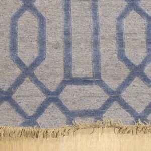 Close-up of a blue and gray flat-woven rug with an interlocking geometric pattern and short fringe on light wood flooring.