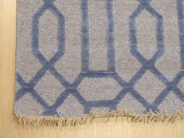 Close-up of a blue and gray flat-woven rug with an interlocking geometric pattern and short fringe on light wood flooring.