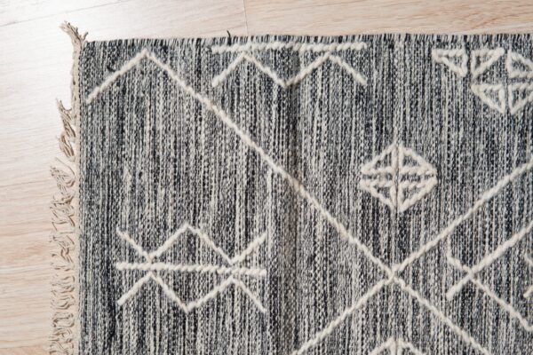 Variegated black and cream rug with thick raised geometric patterns and fringe rests on light wood floor.