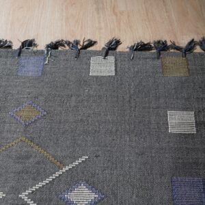 A dark gray flatweave rug with geometric shapes and tied fringes rests on light wood plank flooring.