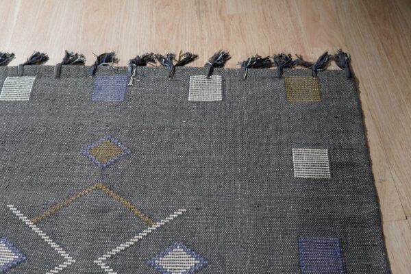 A dark gray flatweave rug with geometric shapes and tied fringes rests on light wood plank flooring.