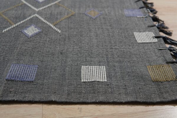 Dark gray flatweave rug with blue, white, and gold geometric patterns on light wood floor.