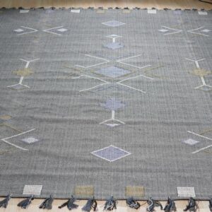 Dark gray flatweave rug with abstract white, blue, and khaki geometric designs and short tassels on wood flooring.