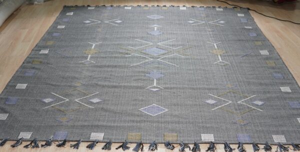 Dark gray flatweave rug with abstract white, blue, and khaki geometric designs and short tassels on wood flooring.