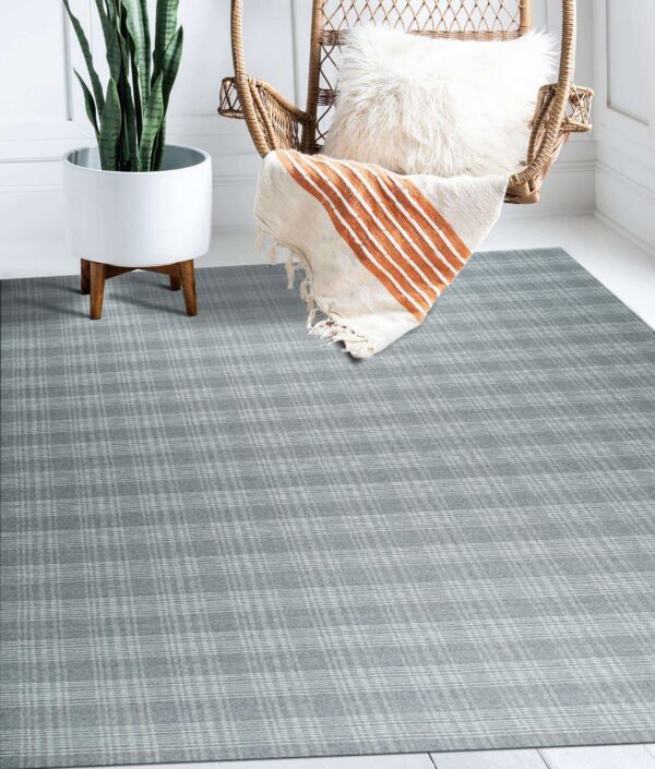 Low pile gray plaid rug covers white wood floors next to a plant and wicker chair.