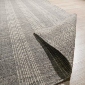 Gray rug with thin vertical stripes partially rolled on light wood flooring.
