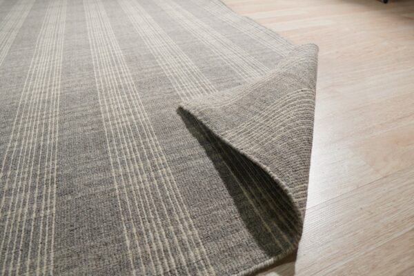 Gray rug with thin vertical stripes partially rolled on light wood flooring.