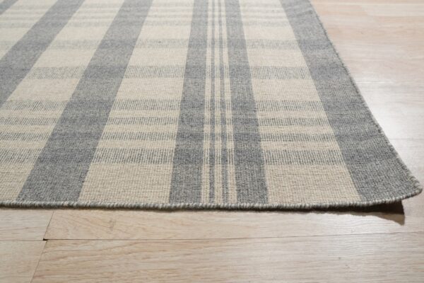 A flatweave rug with a gray and cream plaid pattern rests on light wood flooring.