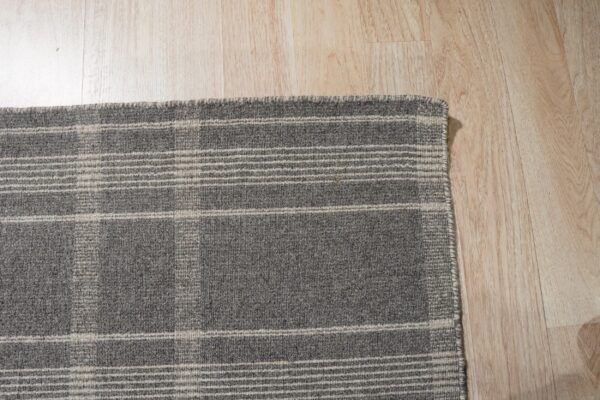 A corner view of a woven gray rug featuring a light beige plaid pattern on blonde flooring.