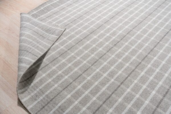 A gray flatwoven rug with a light plaid pattern is partially unrolled on light wood plank flooring.