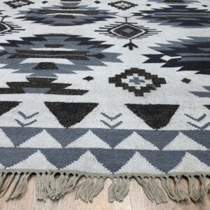 Flat-woven rug with white, gray, and dark charcoal geometric patterns and fringe on light wood flooring.