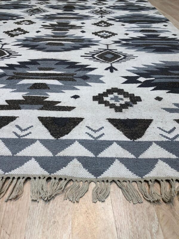 Flat-woven rug with white, gray, and dark charcoal geometric patterns and fringe on light wood flooring.