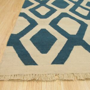 Flatwoven rug with a large teal and cream geometric pattern and visible fringe on light wood floors.
