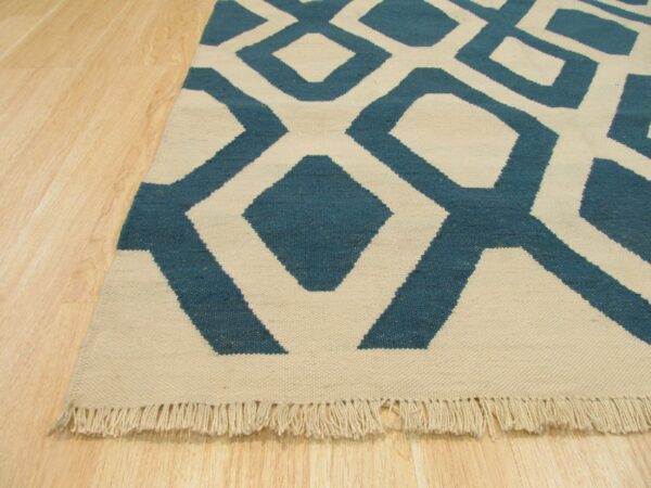 Flatwoven rug with a large teal and cream geometric pattern and visible fringe on light wood floors.