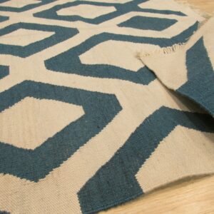 Teal and cream flat-weave rug featuring a geometric pattern is displayed folded on light wood flooring.