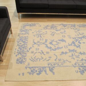 Light beige rug with faded blue traditional patterns sits between a dark sofa and armchair on wood floor.