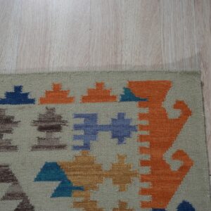 Beige flatweave rug with orange, blue, and brown geometric patterns on pale wood floor.