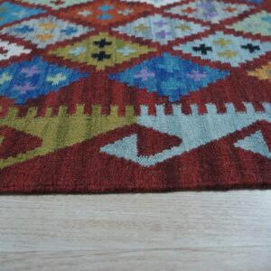 Red flat-weave rug featuring a colorful geometric diamond pattern on light plank flooring.