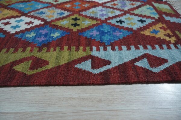 Red flat-weave rug featuring a colorful geometric diamond pattern on light plank flooring.