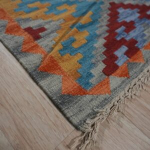 Close-up view of a flat-woven rug with multicolor geometric patterns and fringe on light wood flooring.