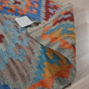 Geometric flatweave rug featuring stepped patterns in blue, orange, gray, and brown on light plank flooring.