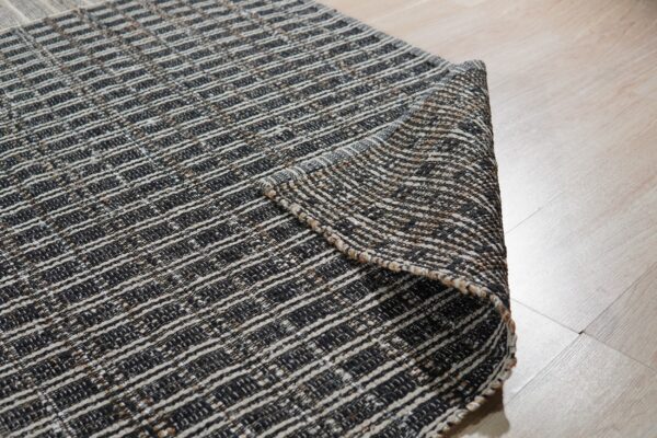 Close-up of a black, white, and tan striped flatweave rug, partially rolled back on light wood flooring.